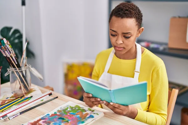 African american woman artist reading book with serious expression at art studio