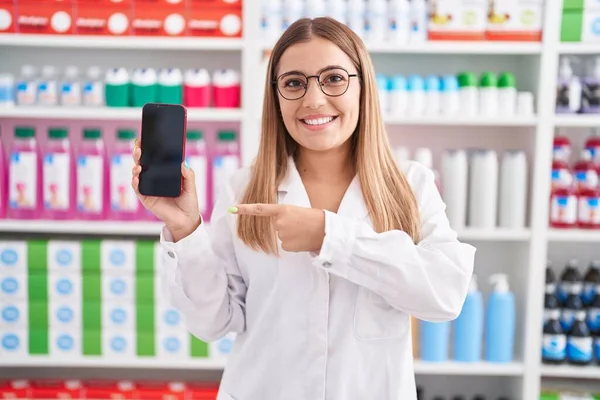 Young Blonde Woman Working Pharmacy Drugstore Showing Smartphone Screen Smiling — Zdjęcie stockowe