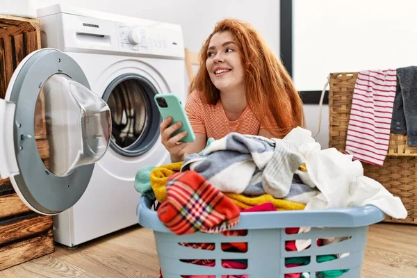 洗濯室でスマートフォンの洗濯服を使用して若い赤毛の女性 — ストック写真