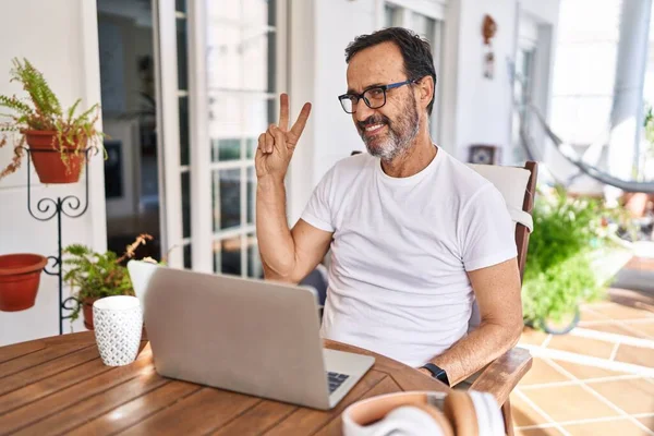 Middle age man using computer laptop at home smiling looking to the camera showing fingers doing victory sign. number two.