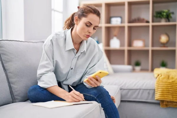 Young Woman Writing Notebook Using Smartphone Home — Foto de Stock