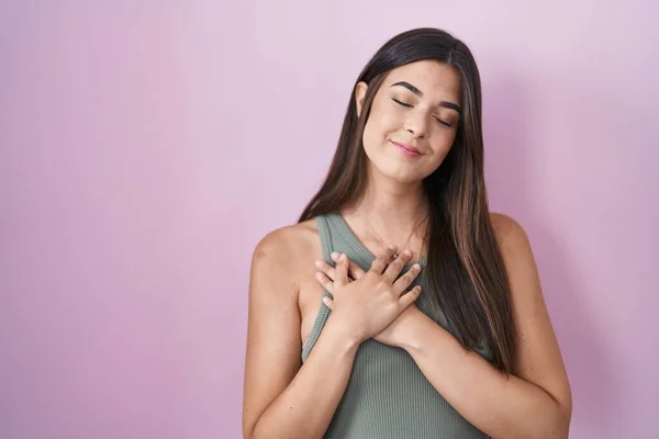 ピンクの背景の上に立つヒスパニック系の女性は 目を閉じて胸に手を笑顔と顔に感謝のジェスチャー 健康の概念 — ストック写真