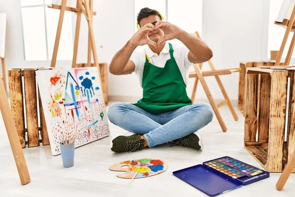 Young hispanic man at art studio smiling in love doing heart symbol shape with hands. romantic concept.
