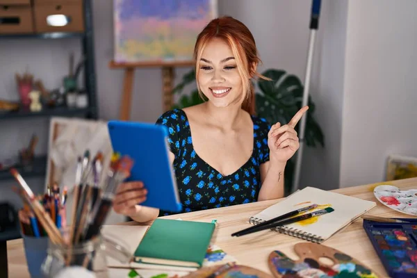 年轻的高加索女人坐在艺术工作室里做视频通话 带着平板电脑的微笑 手指手画脚地指向旁边 — 图库照片