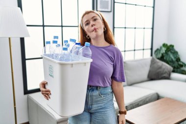 Geri dönüşüm sepetini plastik şişelerle tutan genç kızıl saçlı kadın dudaklarıyla balık suratı yapıyor, çılgınca ve komik bir jest. Komik ifade. 