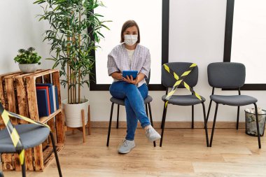 Middle age hispanic woman wearing safety mask using tablet at waiting room