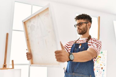 Sanat stüdyosunda tuval tutan, ciddi ifadeli genç bir Arap sanatçı..