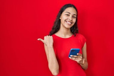 Genç Brezilyalı kadın kırmızı arka planda akıllı telefon kullanıyor. Arkayı işaret ediyor. El ve baş parmakları havada, kendinden emin gülümsüyor. 
