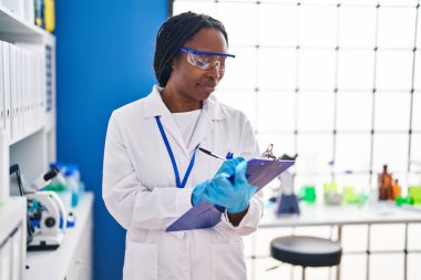 African american woman wearing scientist uniform writing on clipboard laboratory