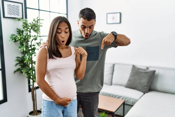 Young interracial couple expecting a baby, touching pregnant belly pointing down with fingers showing advertisement, surprised face and open mouth