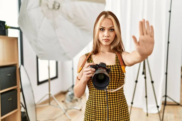 年轻的高加索摄影师女孩手持专业相机在摄影棚用张开的手做停止签名 带着严肃而自信的表情和防御姿态 — 图库照片