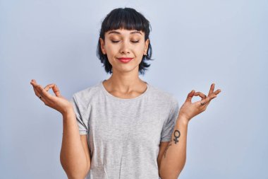 Mavi arka plan üzerine gündelik tişört giyen genç İspanyol kadın rahatla ve parmaklarıyla meditasyon hareketi yaparken gözleri kapalı gülümse. yoga konsepti. 