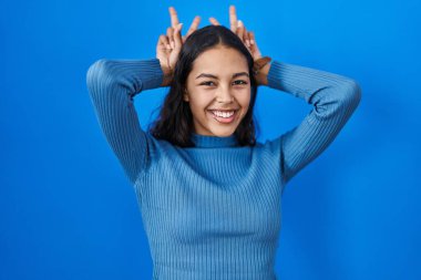 Genç Brezilyalı bir kadın mavi arka planda duruyor. Komik ve çılgın pozlar veriyor. Parmakları kafasında tavşan kulağı gibi, neşeli bir şekilde gülümsüyor. 
