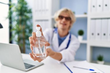 Genç sarışın doktor, klinikte dezenfektan jeli kullanan kendine güvenen gülümseyen bir adam.