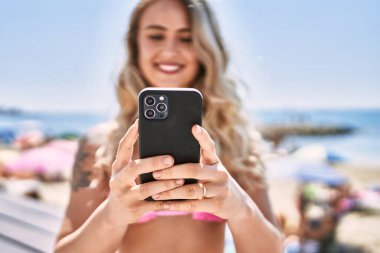 Kumsalda bankta oturan akıllı telefon kullanan genç sarışın kız..