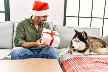 Genç İspanyol bir adam elinde hediyeyle evde köpekle oturuyor.