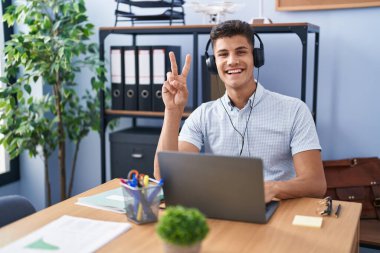 Ofiste çalışan, kulaklık takan, gülümseyen, parmaklarını zafer işareti yaparken gösteren genç İspanyol bir adam. İki numara.. 