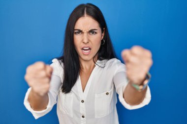Mavi arka planda öfkeli ve kızgın duran genç İspanyol kadın öfkeyle bağırırken kızgın ve öfkeli yumruklar sallıyor. öfke ve agresif konsept. 