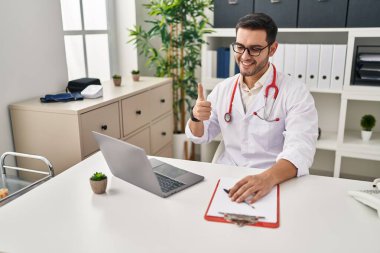 Sakallı genç İspanyol doktor klinikte online randevu veriyor mutlu ve olumlu gülümsüyor, başparmağıyla mükemmel ve onay işareti yapıyor. 