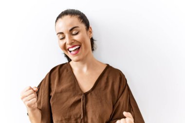 Genç esmer kadın, izole edilmiş arka planda mutlu ve heyecanlı bir şekilde kollarını kaldırıp, gülümseyerek ve başarı çığlıkları atarak zafer jesti yapıyor. kutlama konsepti. 