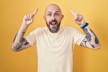 Sarı arka planda dikilen İspanyol dövmeli adam. Şaşırmış, şaşırmış, parmaklarıyla işaret ediyor ve kollarını kaldırıyordu.. 