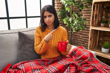 Kanepede oturan Latin asıllı genç bir kadın evde kahve içerken elini kaldırıyor ve reklamını yapıyor, ciddi ve sakin bir yüz ifadesi var. 