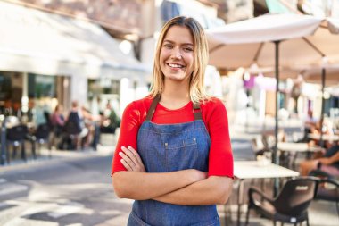 Young hispanic woman waitress standing with arms crossed gesture at coffee shop terrace