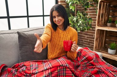 Genç İspanyol kadın kanepede oturup kahve içerken dostça gülümseyerek selamlaşma ve selamlaşma sunuyor. başarılı iş. 