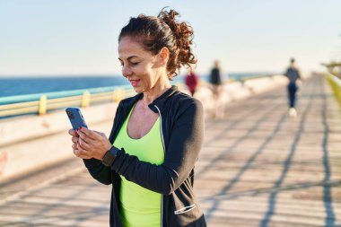 Middle age hispanic woman working out with smartphone at promenade