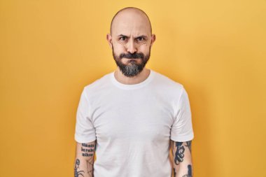 Sarı arka planda dikilen dövmeli, komik yüzlü İspanyol genç adam. Havayla şişmiş ağız, çılgın bir ifade.. 