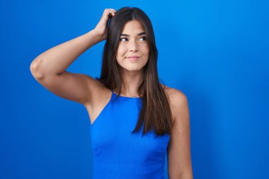 Hispanic woman standing over blue background confuse and wondering about question. uncertain with doubt, thinking with hand on head. pensive concept. 