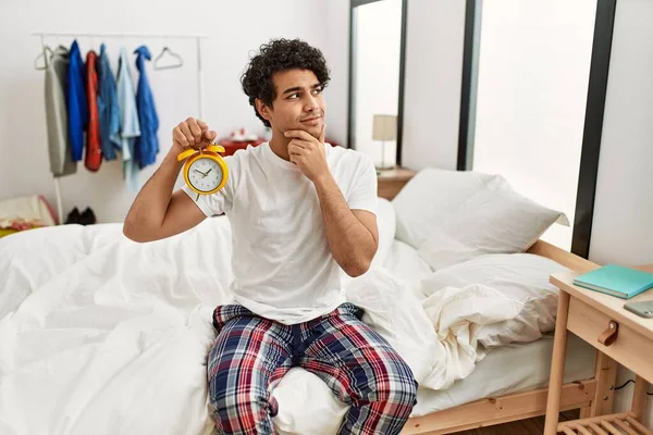 Young hispanic man holding alarm clock sitting on the bedroom serious face thinking about question with hand on chin, thoughtful about confusing idea