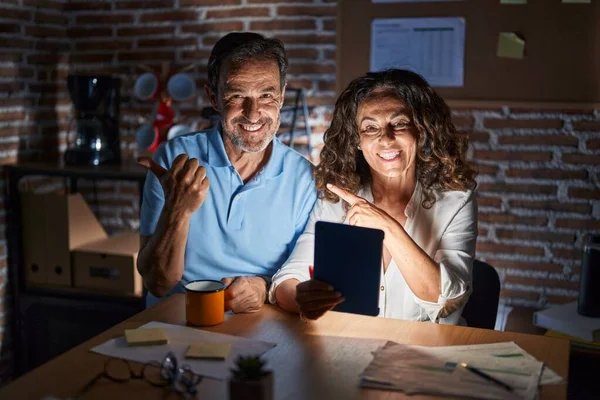 Middle age hispanic couple using touchpad sitting on the table at night pointing to the back behind with hand and thumbs up, smiling confident