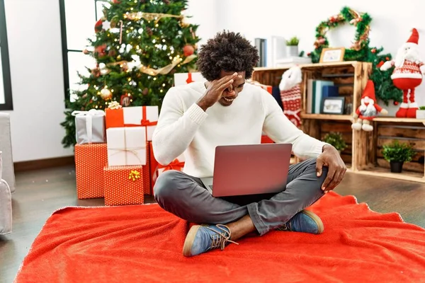 Genç Afro-Amerikalı adam dizüstü bilgisayar kullanıyor Noel ağacının yanında oturmuş gülümsüyor ve gülümsüyor. 
