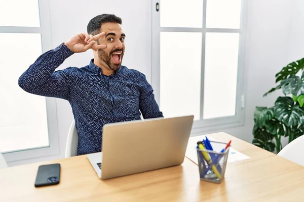 顔の上に指で平和のシンボルを行うラップトップでオフィスで働くひげを持つ若いヒスパニック系の男 勝利を示す明るい笑顔 — ストック写真