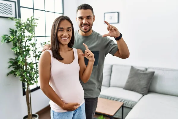 Young interracial couple expecting a baby, touching pregnant belly smiling and confident gesturing with hand doing small size sign with fingers looking and the camera. measure concept.