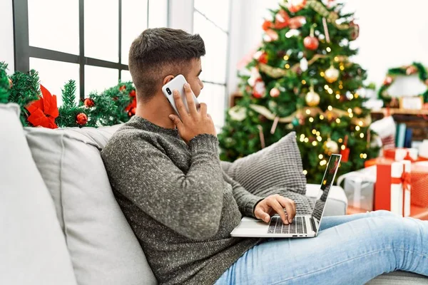年轻的他的惊慌失措的男人在家里坐在圣诞树旁的沙发上 用笔记本电脑和智能手机聊天 — 图库照片