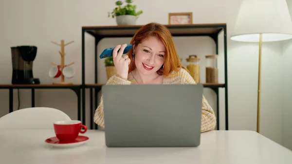 年轻的红头发女人在家用智能手机用笔记本电脑听音频讯息 — 图库照片