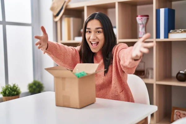 Young south asian woman opening cardboard box looking at the camera smiling with open arms for hug. cheerful expression embracing happiness.