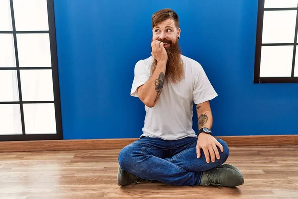 Redhead man with long beard sitting on the floor at empty room looking stressed and nervous with hands on mouth biting nails. anxiety problem.