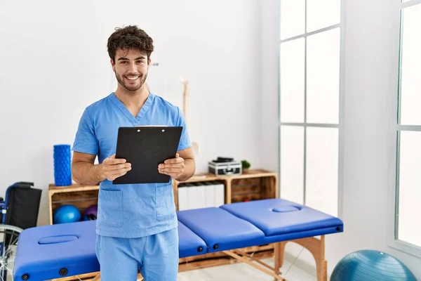 Young Hispanic Man Wearing Physio Therapist Uniform Holding Clipboard Clinic — Stockfoto