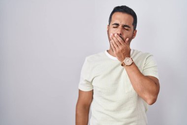 Hispanic man with beard standing over isolated background bored yawning tired covering mouth with hand. restless and sleepiness. 