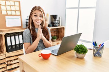 Genç esmer kadın ofiste dizüstü bilgisayarla çalışıyor birlikte dua edip af diliyor kendinden emin bir şekilde gülümsüyor.. 