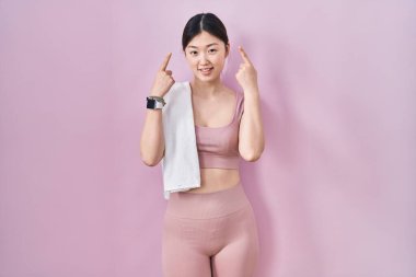 Spor kıyafetleri ve havlu giymiş Çinli genç bir kadın iki eliyle başını işaret ederek gülümsüyor. Harika bir fikir ya da düşünce, iyi bir hafıza. 