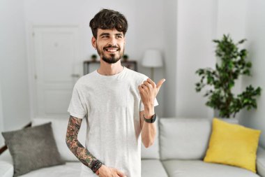 Oturma odasında sakallı, mutlu yüzüyle gülümseyen ve baş parmağıyla yüzünü gösteren İspanyol bir adam.. 