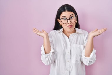 Pembe arka planda duran esmer bir kadın. Kolları ve elleri havada, kafası karışmış bir ifade. şüphe kavramı. 