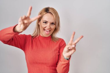 Sarışın kadın, izole edilmiş arka planda dikiliyor, gülümsüyor ve parmaklarını iki eliyle zafer işareti yapıyor. İki numara.. 