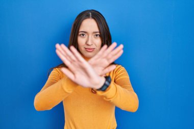 Genç esmer kadın mavi arka planda duruyor. Reddedilme ifadesi. Kollar ve avuçlar çapraz. 