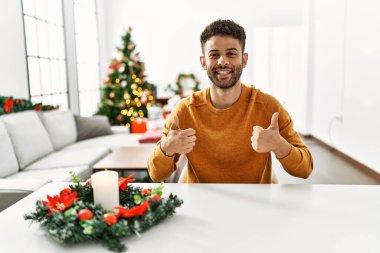 Arap genç adam Noel ağacının yanında masada oturuyor. Elleriyle olumlu jest yapıyor. Gülümsüyor ve mutlu. Neşeli ifade ve kazanan jest. 