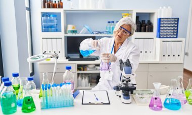 Middle age woman wearing scientist uniform pouring liquid on test tube at laboratory